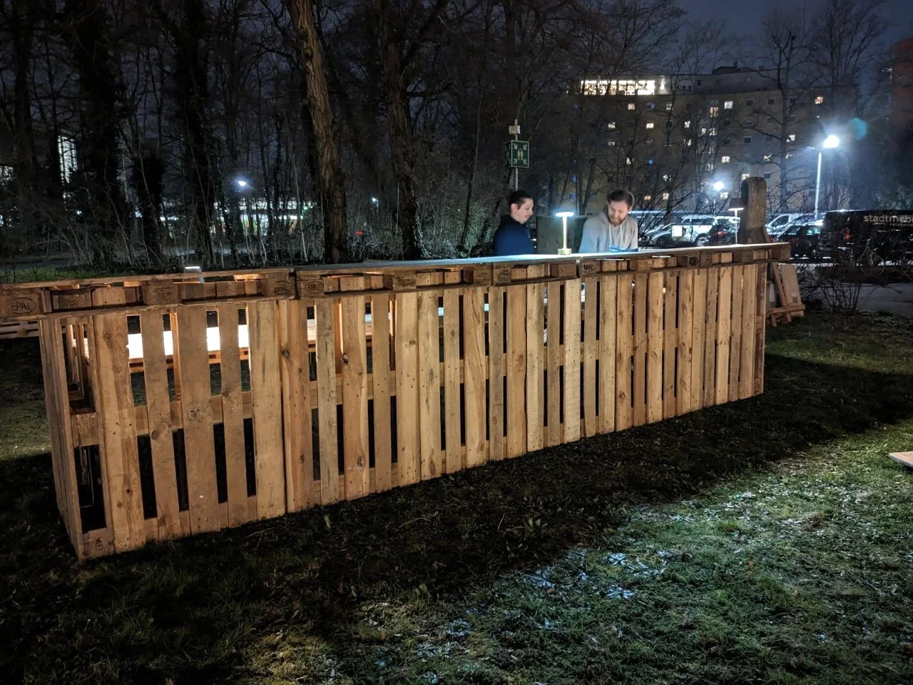 Die mobile Bar bei Nacht: warmes Licht scheint durch die Paletten, zwei Menschen stehen dahinter.