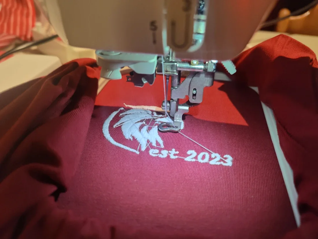 Detailaufnahme einer Stickmaschine, die das Kupfer-Adler-Logo auf bordeauxroten Stoff stickt.